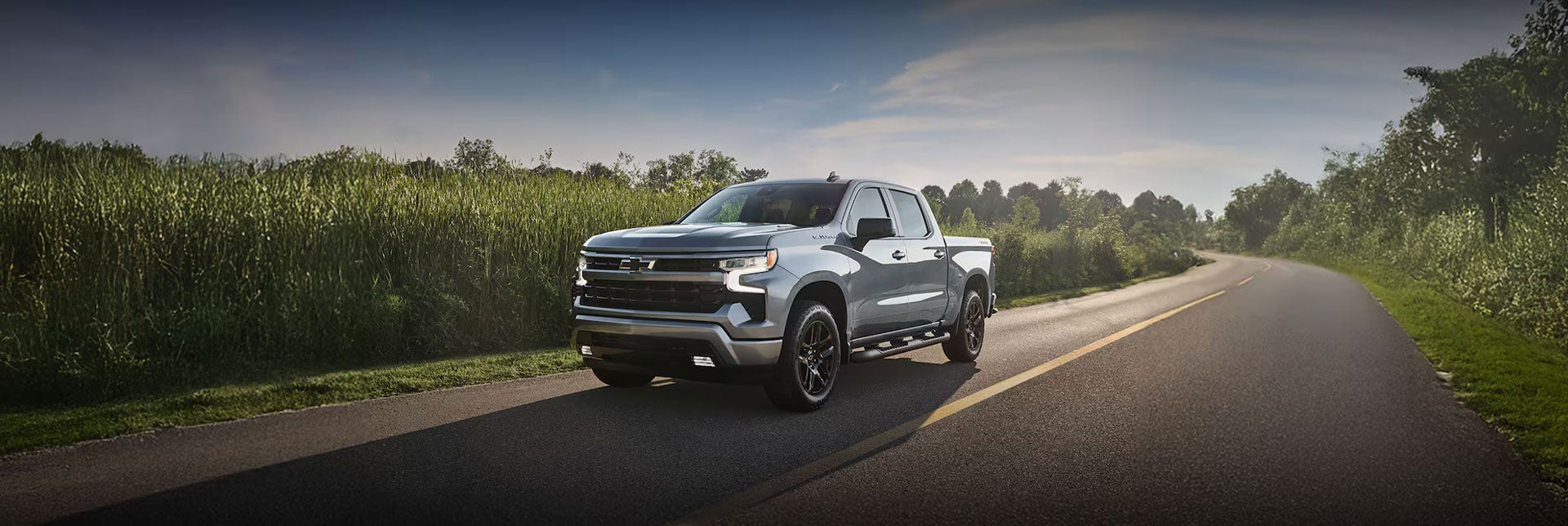 Three-quarter View of a Chevy Silverado 1500 Driving Down a Road Lined with Green Foliage with Text Overlay Reading "Can't wait?"